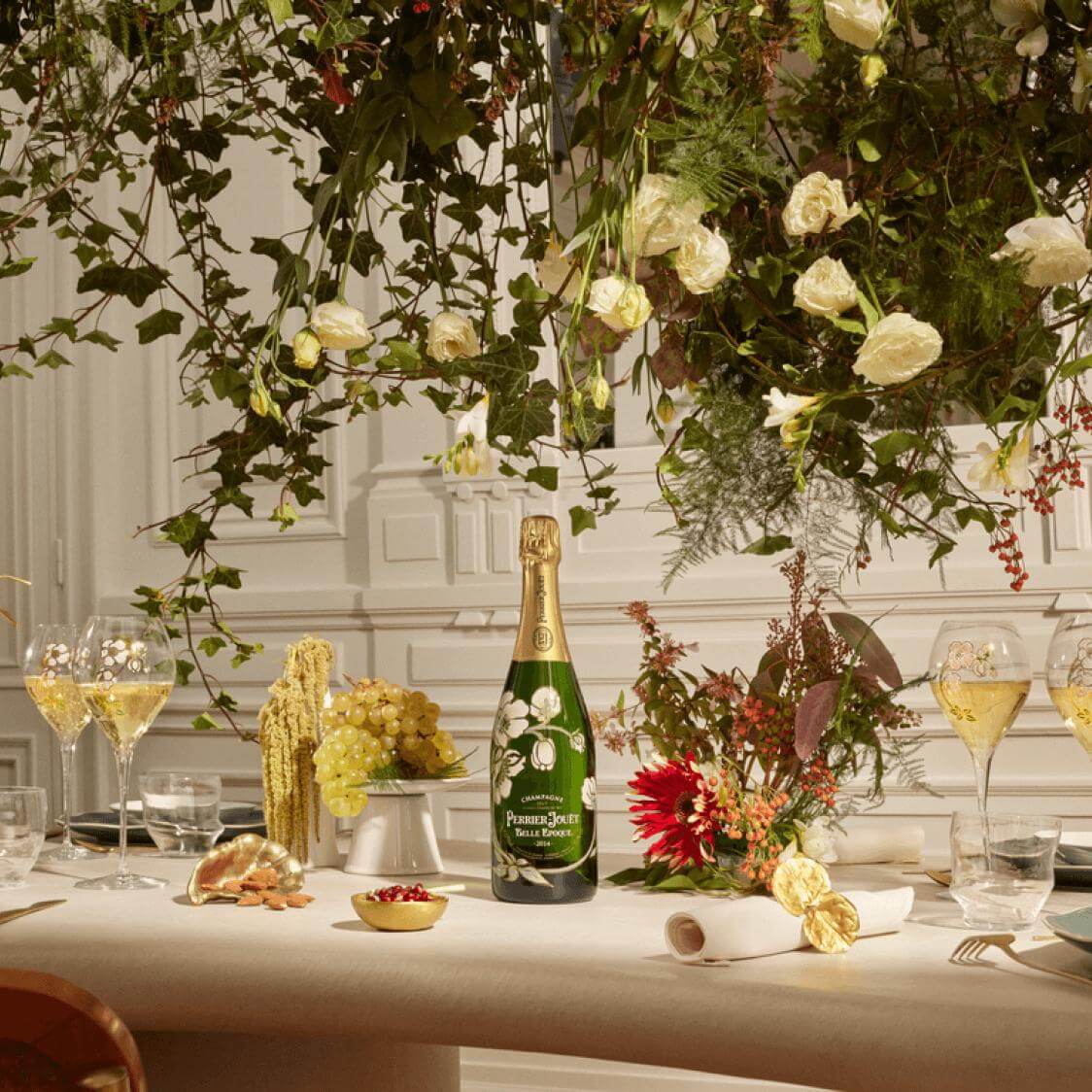 Perrier-Jouet Grand Brut Champagne bottle on a table with flowers, grapes, and two glasses of champagne