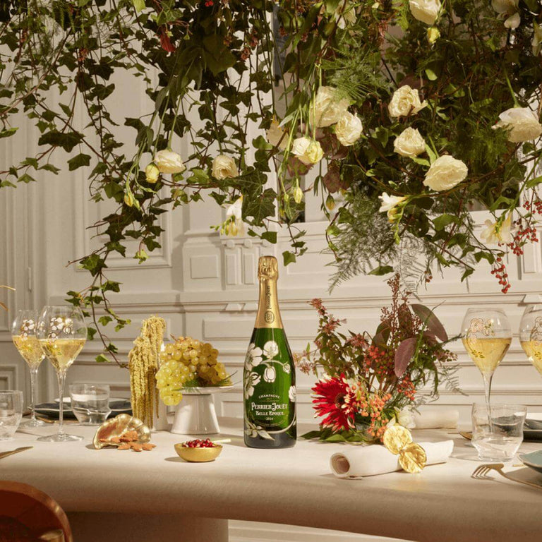 Perrier-Jouet Grand Brut Champagne bottle on a table with flowers, grapes, and two glasses of champagne